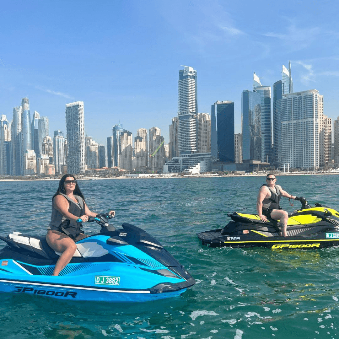 Two riders on jet skis near the Dubai Marina waterfront, with modern high-rise towers in the background on a sunny day