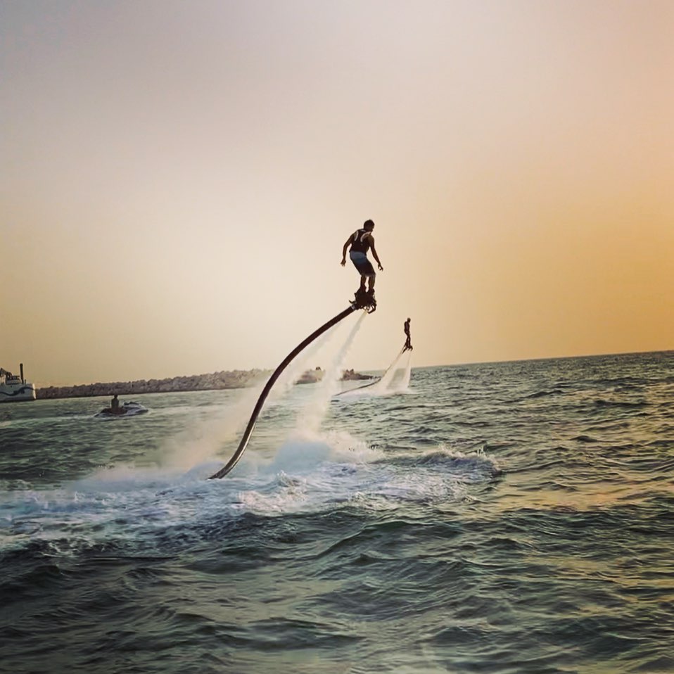 Person flyboarding over the sea in Dubai at sunset, creating a high arc of water with the city skyline in the background.